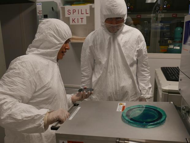 2 scientists in cleanroom garb talking over machinery